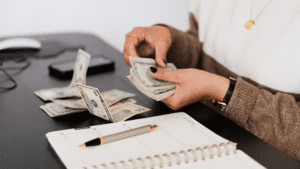 Person counting British currency notes at a desk in the United Kingdom, reviewing cash flow and financial records in a notebook as part of business finance planning.